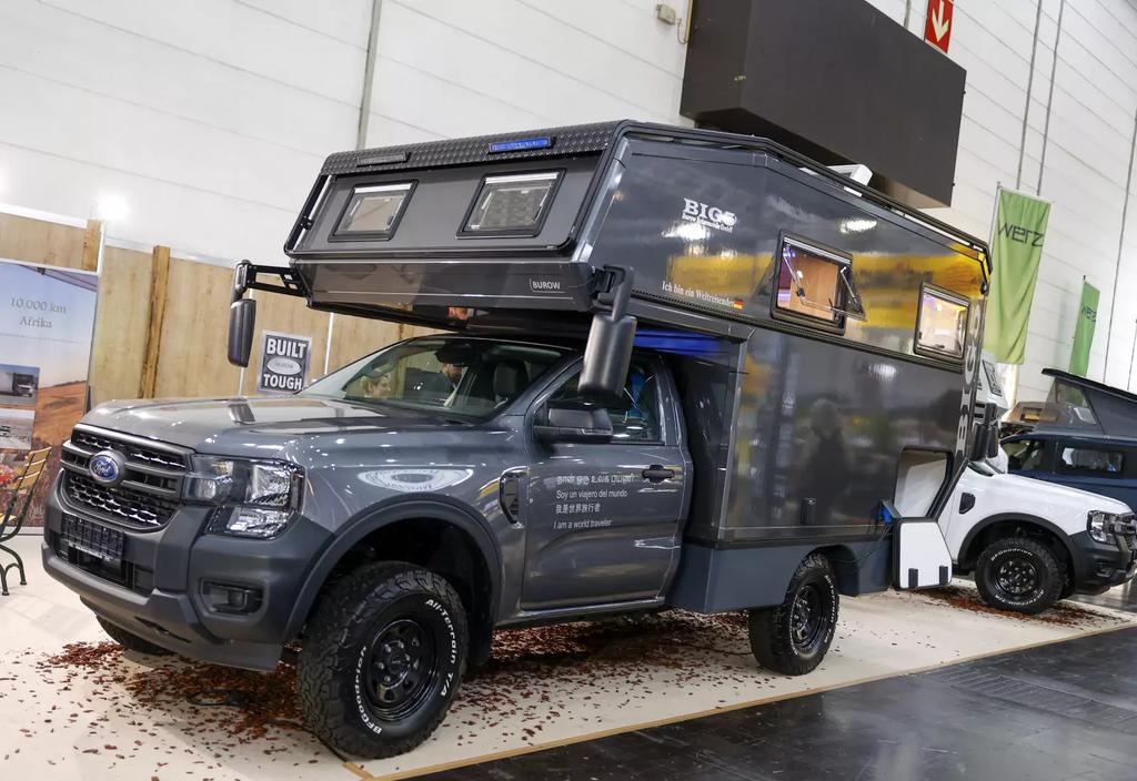 Grauer Pickup mit aufgesetzter erh&ouml;hter Wohnkabine in einer Messehalle; rechts teilweise ein wei&szlig;es Fahrzeug sichtbar