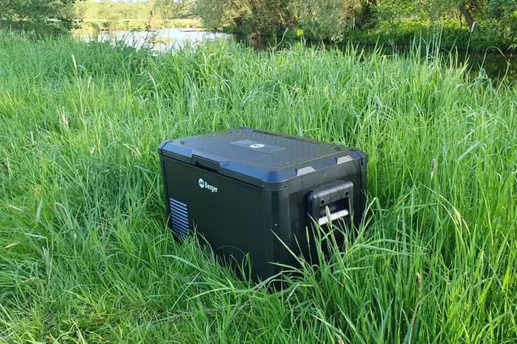 Schwarzes rechteckiges tragbares Gerät mit Tragegriff und Lüftungsschlitzen steht im hohen Gras an einem Ufer; im Hintergrund Wasser und Bäume