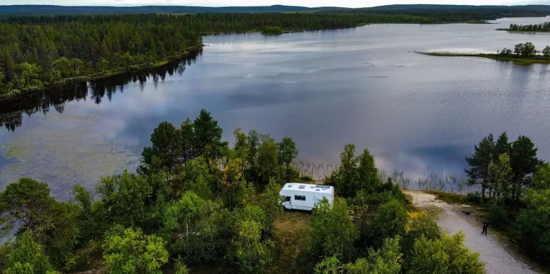 Wei&szlig;es Wohnmobil zwischen B&auml;umen an einem Seeufer mit Sandstrand und Wald im Hintergrund