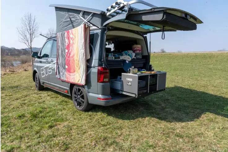 Grauer Camper mit ge&ouml;ffneter Heckklappe auf einer Wiese, herausgezogene Schubladen mit Campingausr&uuml;stung, Handtuch h&auml;ngt an der T&uuml;r, Kissen und Decke im Innenraum sichtbar