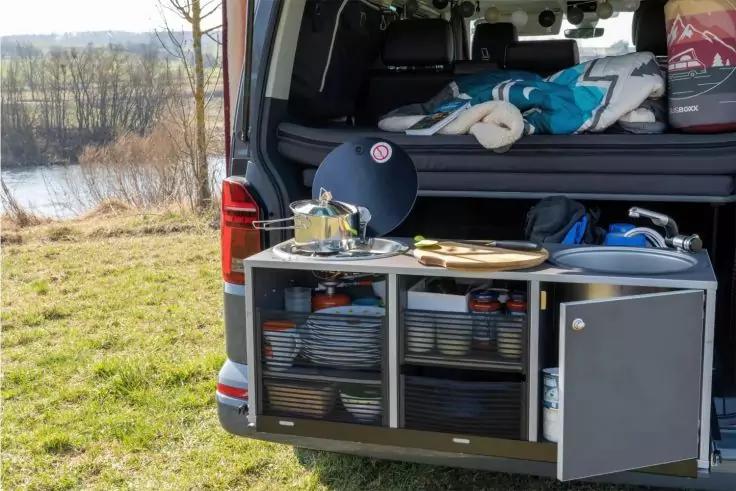 Ge&ouml;ffnete Heckt&uuml;r eines Vans mit ausgezogener K&uuml;chenbox: Kochgeschirr (Topf auf Herd), Schneidebrett, gestapelte Teller und Becher, offenes Seitenfach mit Zylinder, Decken und Kissen im Innenraum, Wiese und Fluss im Hintergrund