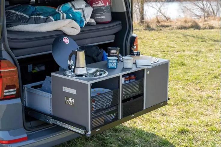 Offener Kofferraum eines Vans mit ausziehbarem Schrankmodul; auf der Arbeitsfl&auml;che eine Metallkanne, Becher, Teller und Aufbewahrungsboxen; Regalf&auml;cher mit Geschirr; im Hintergrund Wiese und Gew&auml;sser