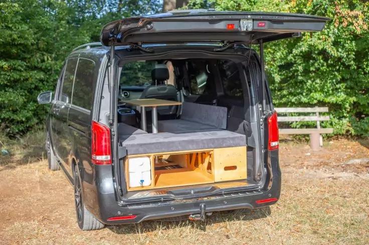 Schwarzes Fahrzeug mit ge&ouml;ffneter Heckklappe; im Laderaum eine Liegefl&auml;che, ein kleiner Tisch und helle Holzschubladen; im Hintergrund B&auml;ume und ein Zaun