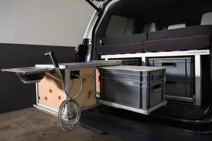 Offenes Heck eines Fahrzeugs mit ausziehbarem Metallschubladensystem; links ein h&ouml;lzernes Schubfach mit rundem Becken, verchromtem Wasserhahn und aufgewickeltem Schlauch; rechts eine graue Kunststoffbox auf einer Schublade; Sitze/Kissen im Hintergrun