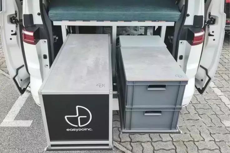 Offenes Heck eines wei&szlig;en Transporters mit zwei herausgezogenen, rechteckigen Lagerkisten: links ein schwarzer Kasten mit wei&szlig;em Logo, rechts zwei graue Schubladen; gepflasterter Parkplatz sichtbar