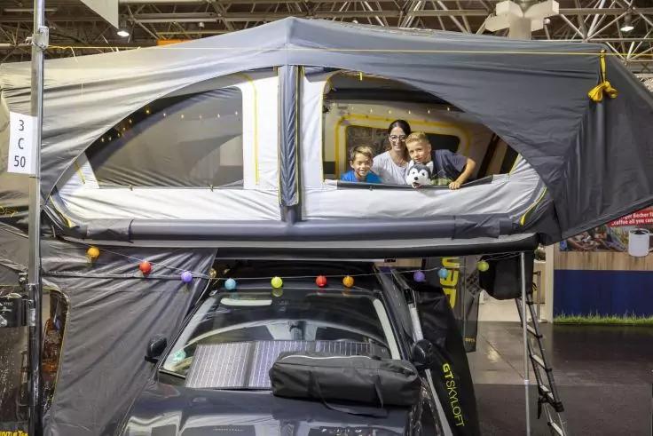 Dachzelt auf einem Auto in einer Ausstellungshalle, durch ein Seitenfenster sind drei Personen sichtbar, Lichterkette an der Vorderseite des Zelts