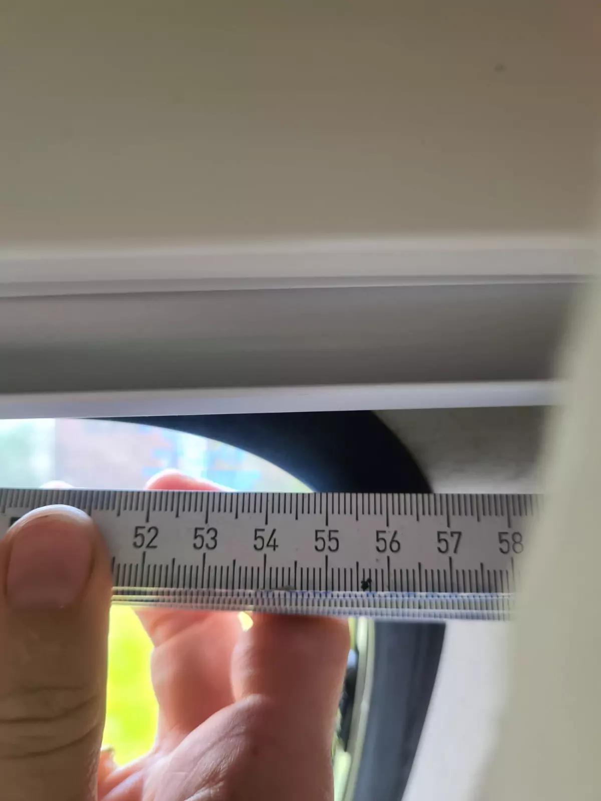 Hand h&auml;lt einen Metallma&szlig;stab waagerecht an eine schmale &Ouml;ffnung zwischen hellen Kanten; Ma&szlig;stabsmarkierungen 52&ndash;58 mm sichtbar; durch die &Ouml;ffnung unscharfe gr&uuml;ne Vegetation und ein roter Fleck sichtbar