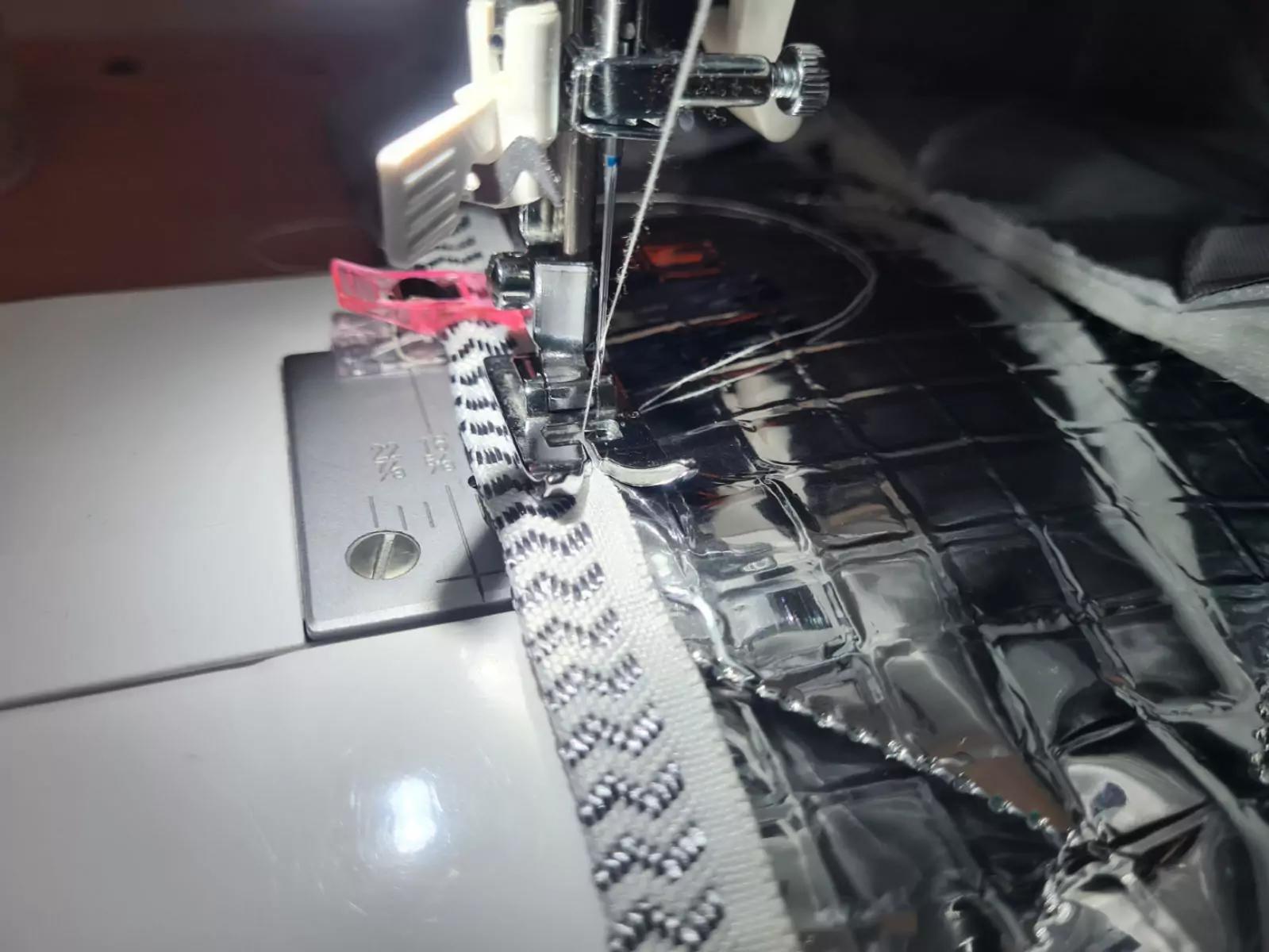 N&auml;hfu&szlig; und Nadel einer N&auml;hmaschine &uuml;ber einem Rei&szlig;verschluss neben gl&auml;nzend reflektierendem Stoff