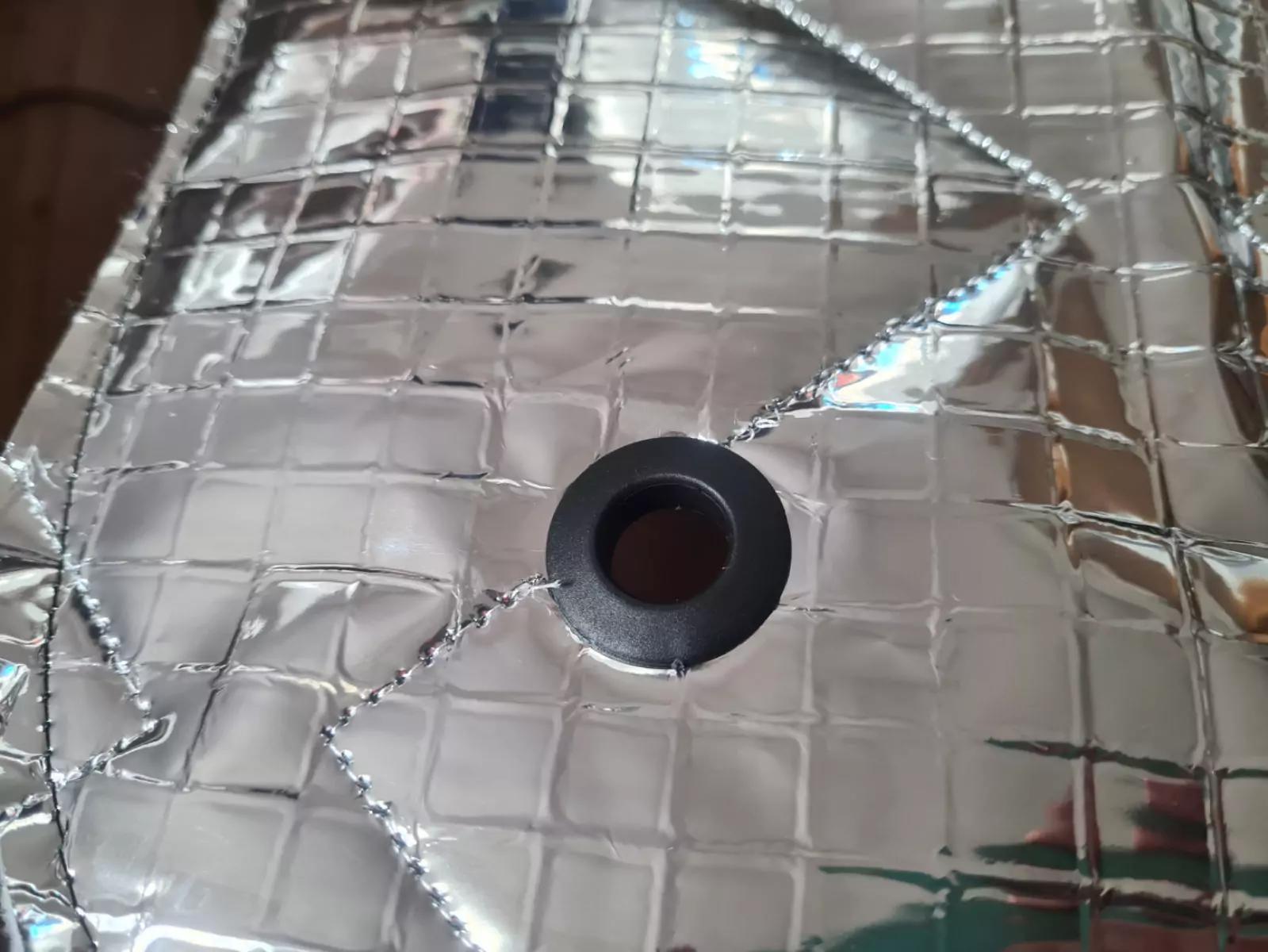 Silbernes, gestepptes, reflektierendes Material mit quadratischem Steppmuster und schwarzer Kunststoff-&Ouml;se in der Bildmitte; oben links Holzfl&auml;che sichtbar