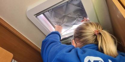 Person von hinten mit blondem Pferdeschwanz in blauem Sweatshirt greift in eine ge&ouml;ffnete rechteckige Dachluke mit silberner Isolierfolie; Holzwand rechts, gemustertes Sitzpolster links