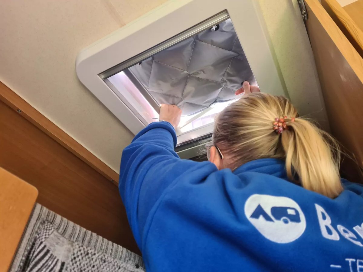 Person von hinten mit blondem Pferdeschwanz in blauem Sweatshirt greift in eine ge&ouml;ffnete rechteckige Dachluke mit silberner Isolierfolie; Holzwand rechts, gemustertes Sitzpolster links