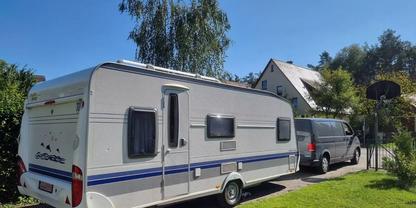 Wei&szlig;er Wohnwagen an grauem Van angeh&auml;ngt auf Einfahrt neben Rasen, B&auml;umen und H&auml;usern unter blauem Himmel