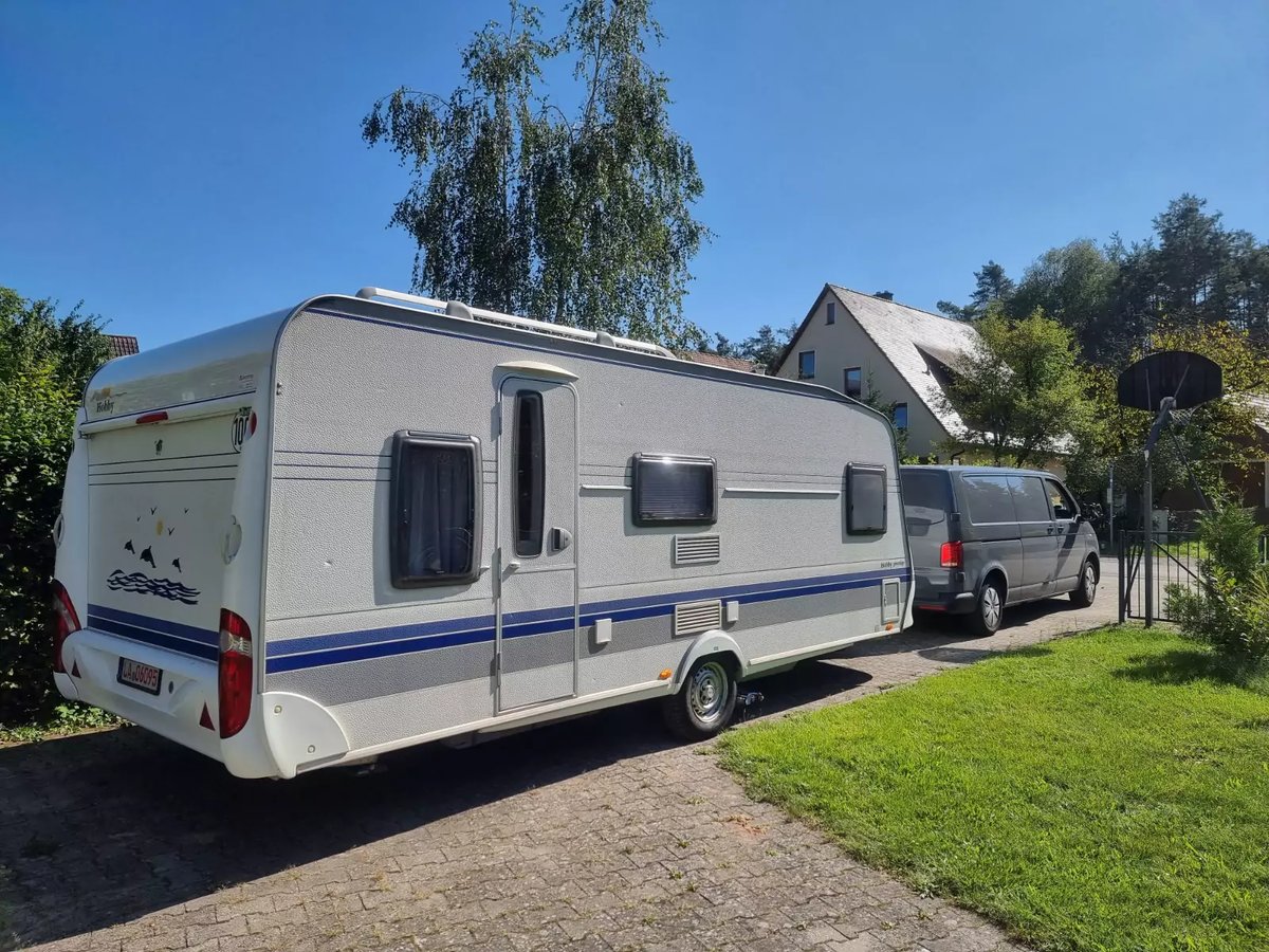 Wei&szlig;er Wohnwagen an grauem Van angeh&auml;ngt auf Einfahrt neben Rasen, B&auml;umen und H&auml;usern unter blauem Himmel