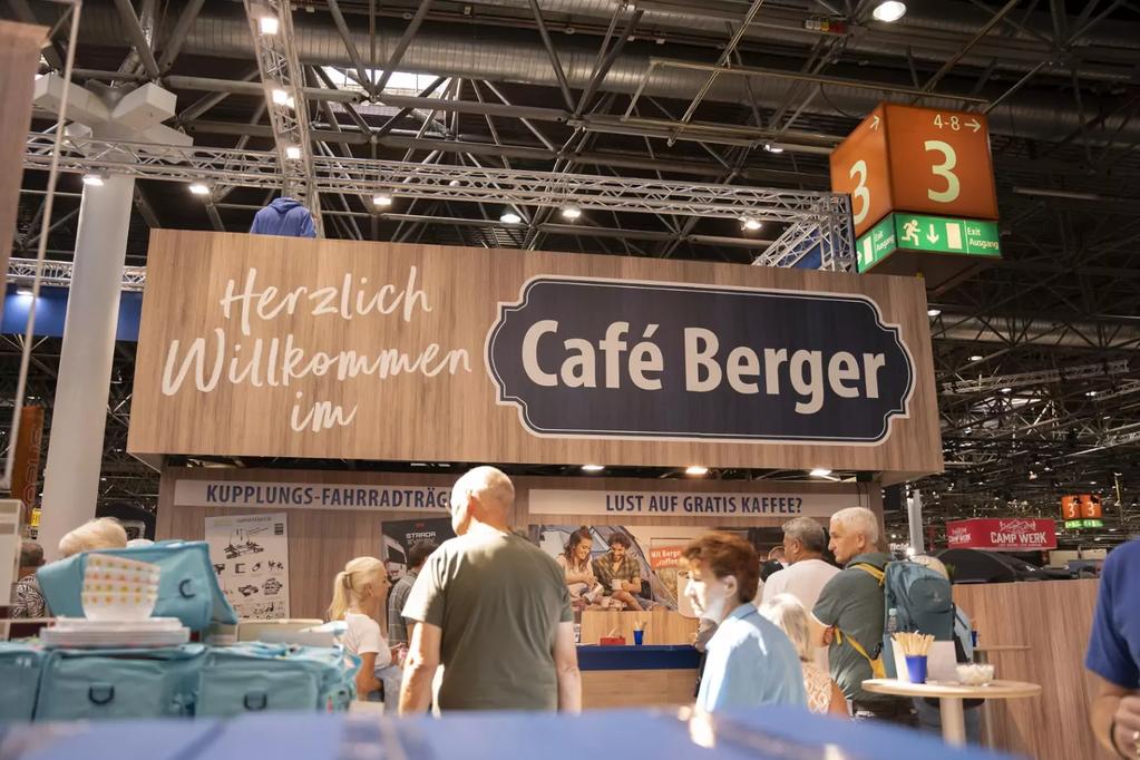 Verkaufsstand mit braunem Schild 'Herzlich Willkommen im Caf&eacute; Berger', mehrere Personen davor, Hallendecke mit Beleuchtung und gr&uuml;nes Schild '3'