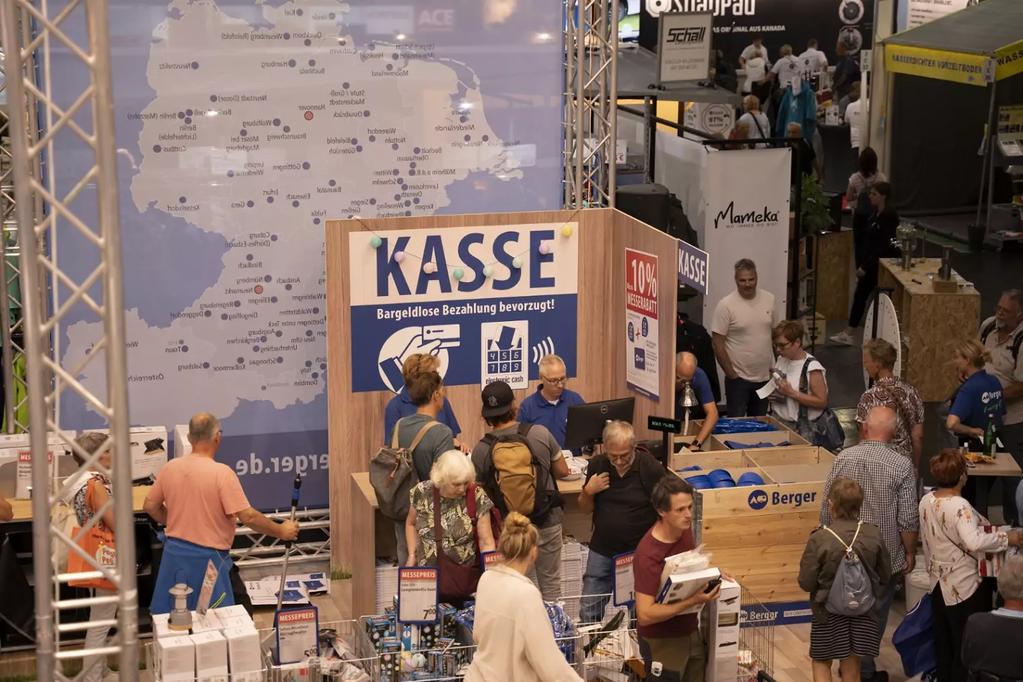 Wand mit gro&szlig;er Karte, davor h&ouml;lzerner Stand mit blauem Schild 'KASSE', mehrere Personen stehen und gehen, Kartons und Brosch&uuml;ren sichtbar