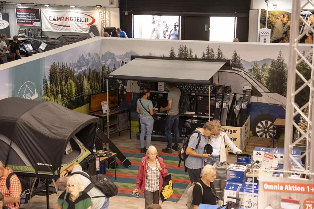 Innenaufnahme eines Ausstellungsstands mit grauem Campinganh&auml;nger und Vordach, zwei M&auml;nner an einem Tresen, mehrere Besucher im Vordergrund