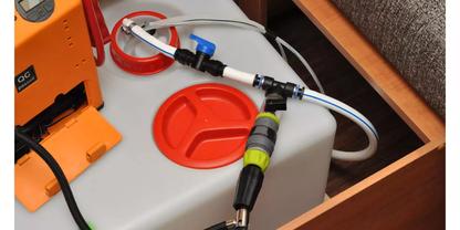 Grauer Kunststoffbeh&auml;lter mit zwei roten Schraubdeckeln in einer Holzschublade, wei&szlig;er Schlauch an schwarzem und gr&uuml;nem Steckverbinder, schwarzes Kabel, teilweise sichtbares oranges Ger&auml;t, grauer Polsterstoff oben