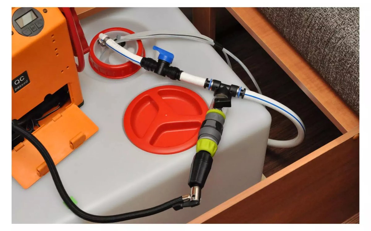 Grauer Kunststoffbeh&auml;lter mit zwei roten Schraubdeckeln in einer Holzschublade, wei&szlig;er Schlauch an schwarzem und gr&uuml;nem Steckverbinder, schwarzes Kabel, teilweise sichtbares oranges Ger&auml;t, grauer Polsterstoff oben