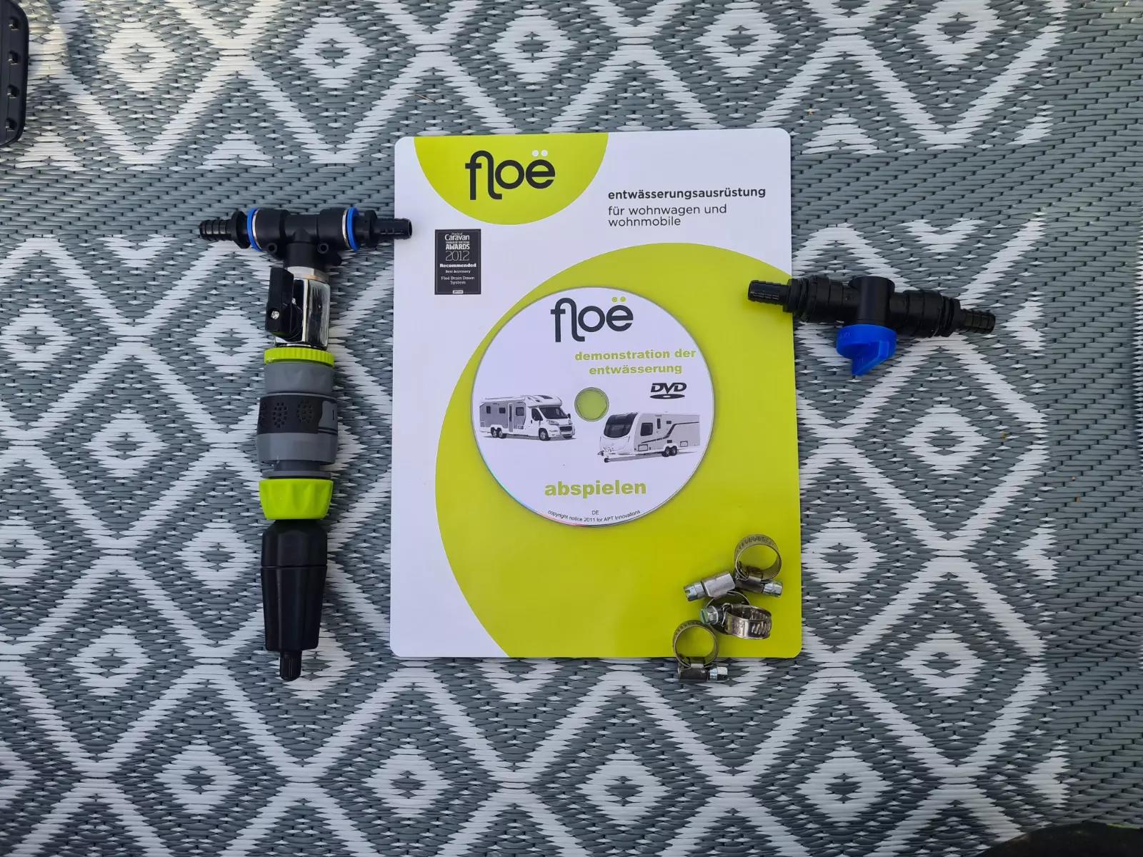 Grauer Teppich mit wei&szlig;em Rautenmuster; darauf eine wei&szlig;-limettengr&uuml;ne Produktverpackung mit runder Aufkleber 'floe'; links ein schwarzes-gr&uuml;nes D&uuml;sen-/Steckteil; rechts ein schwarzer Verbinder mit blauem Ventil; unten rechts drei Metall-Schlauchsche