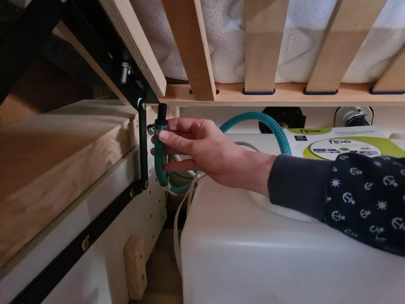 Hand an einem schwarzen Ventil, das an einem gr&uuml;nen Schlauch angeschlossen ist; im Vordergrund ein wei&szlig;er Kunststoffbeh&auml;lter; dar&uuml;ber Holzlatten eines Bettrahmens und eine Metallhalterung