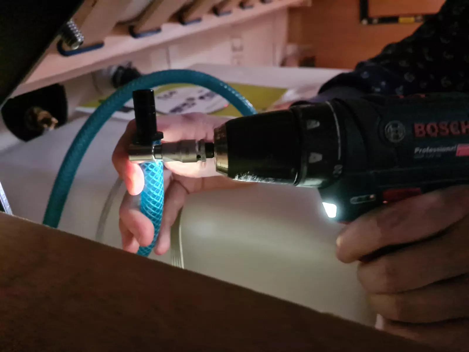 Zwei H&auml;nde halten einen schwarzen Bosch-Akkuschrauber und ein blaues, geriffeltes Kunststoffrohr an einem wei&szlig;en Beh&auml;lter