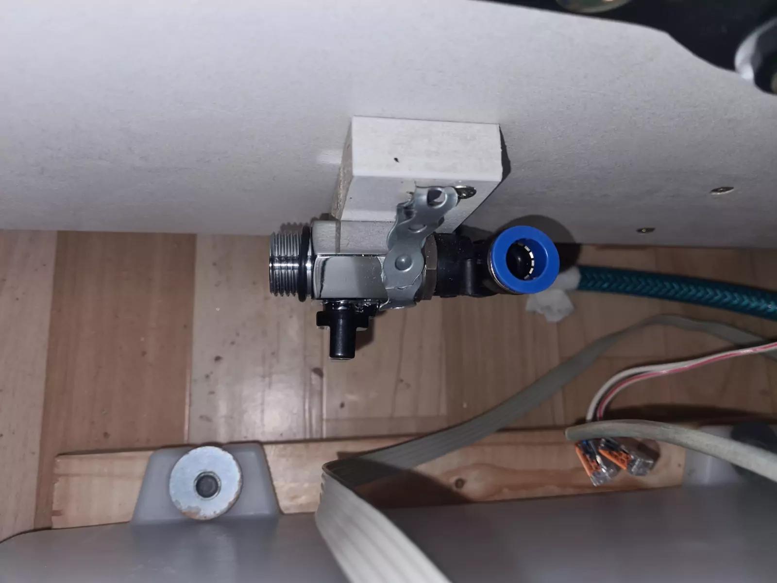 Wei&szlig;e Platte mit montiertem Metallventil und Hebel, blauem Schlauchanschluss, gr&uuml;nem flexiblem Schlauch, Holzunterbau, mehreren Kabeln und einem wei&szlig;en Kunststoffring