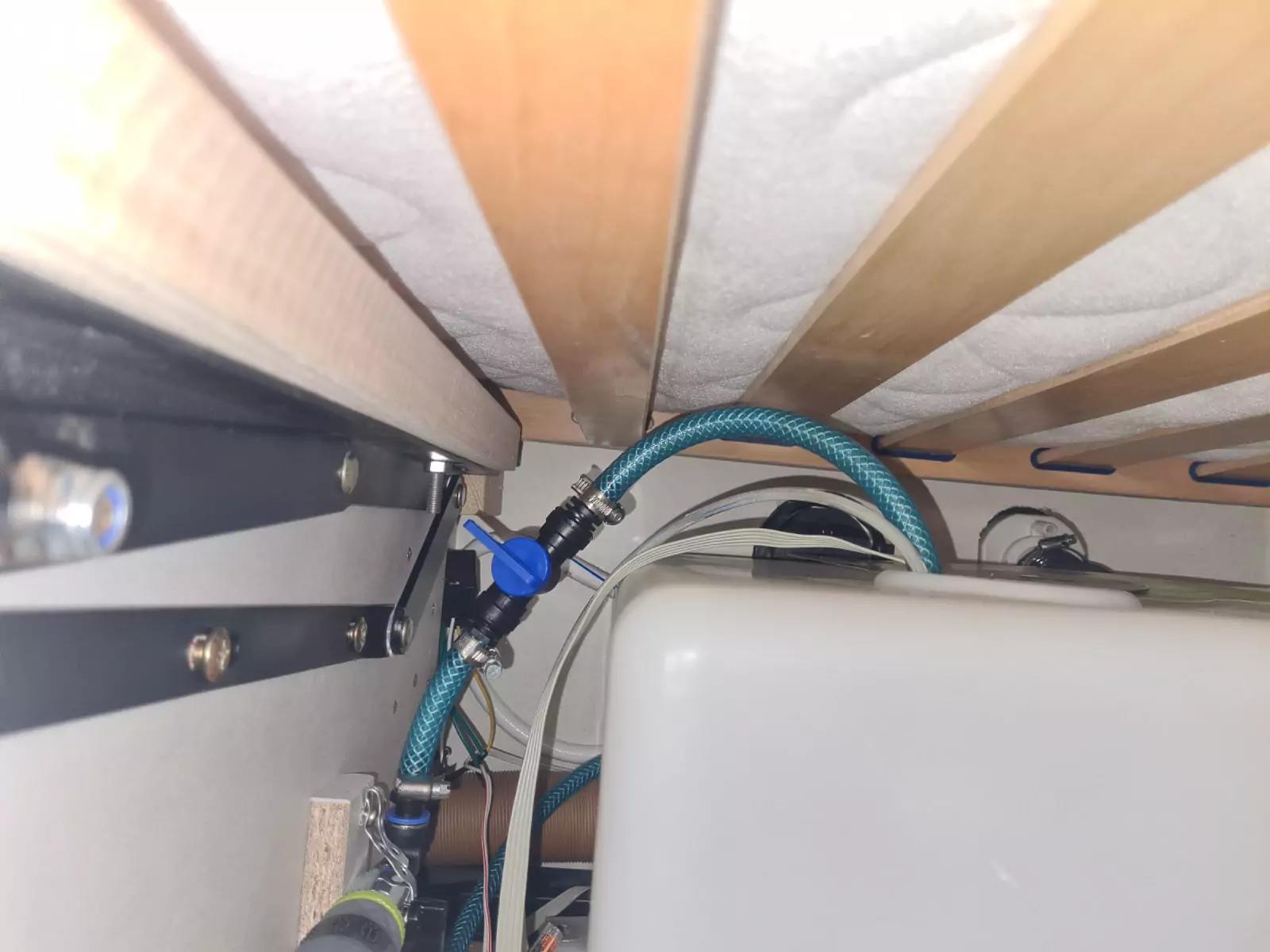 Unterseite einer Matratze mit Holzlatten, ein gr&uuml;n-blauer flexibler Schlauch mit Metallanschl&uuml;ssen, wei&szlig;e Plastikbox, Metallrahmen und Kabel