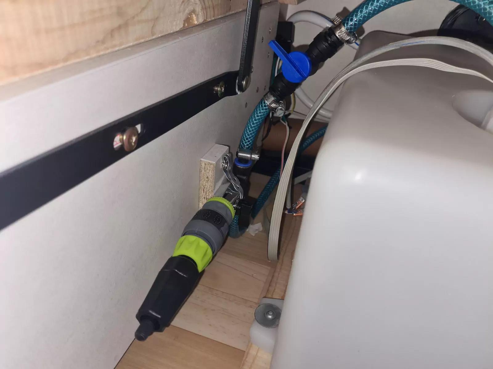 Wei&szlig;er Kunststoffbeh&auml;lter rechts, blau-gr&uuml;ne Gewebeschl&auml;uche und schwarzer Steckverbinder, helles Holzbrett, schwarze Metallleiste mit Schrauben