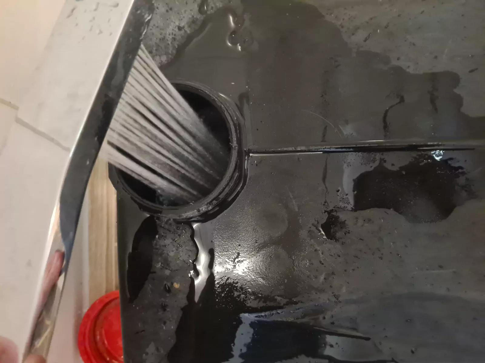 Schwarzer Kunststoffbeh&auml;lter mit Wasserpf&uuml;tzen; wei&szlig;es geripptes Rohr f&uuml;hrt in eine runde &Ouml;ffnung; links ein Chromrand und unten links ein rotes rundes Objekt