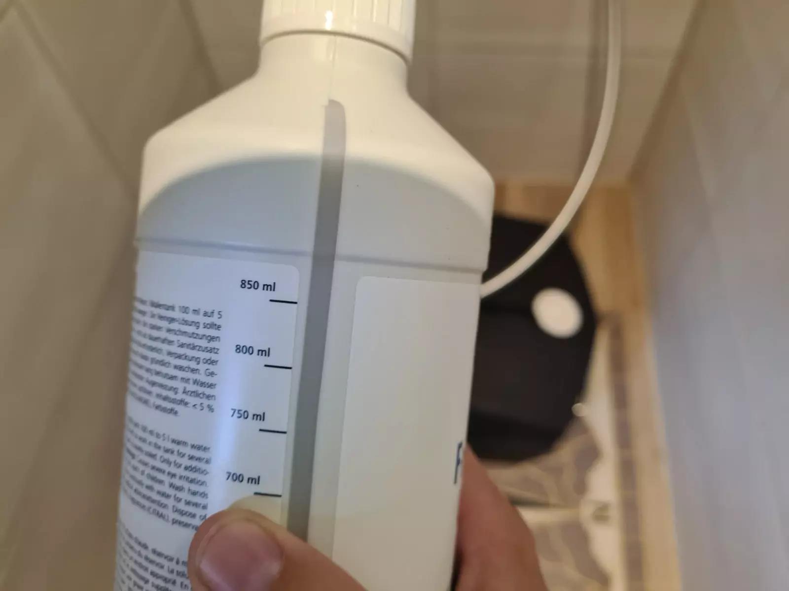 Nahaufnahme einer wei&szlig;en Kunststoffflasche mit vertikaler Milliliter-Skala, von einer Hand gehalten; unscharfer Hintergrund mit Fliesen und einem schwarzen Gegenstand mit wei&szlig;er runder &Ouml;ffnung