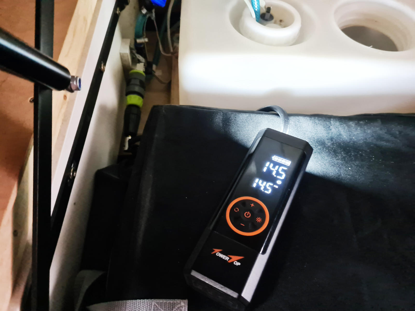 Schwarzes rechteckiges Handger&auml;t mit beleuchtetem Display und orangefarbenem Bedienring auf einer schwarzen Polsterfl&auml;che; rechts wei&szlig;e Kunststofftanks mit runden &Ouml;ffnungen und blauem Verschluss; Schl&auml;uche und Rohrverbindungen im Hintergrund