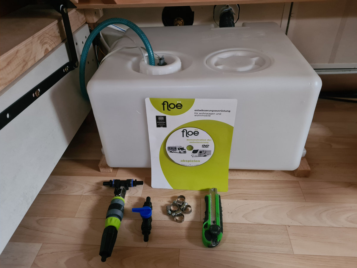 Wei&szlig;er rechteckiger Kunststofftank mit zwei &Ouml;ffnungen oben, gr&uuml;ner Schlauch in einer &Ouml;ffnung; gelb-gr&uuml;nes Heft mit Aufdruck 'floe' davor; Schrauben, Schlauchklemme und mehrere Werkzeuge (Schraubendreher und weiteres Handwerkzeug) auf Holzfu&szlig;boden; Ho