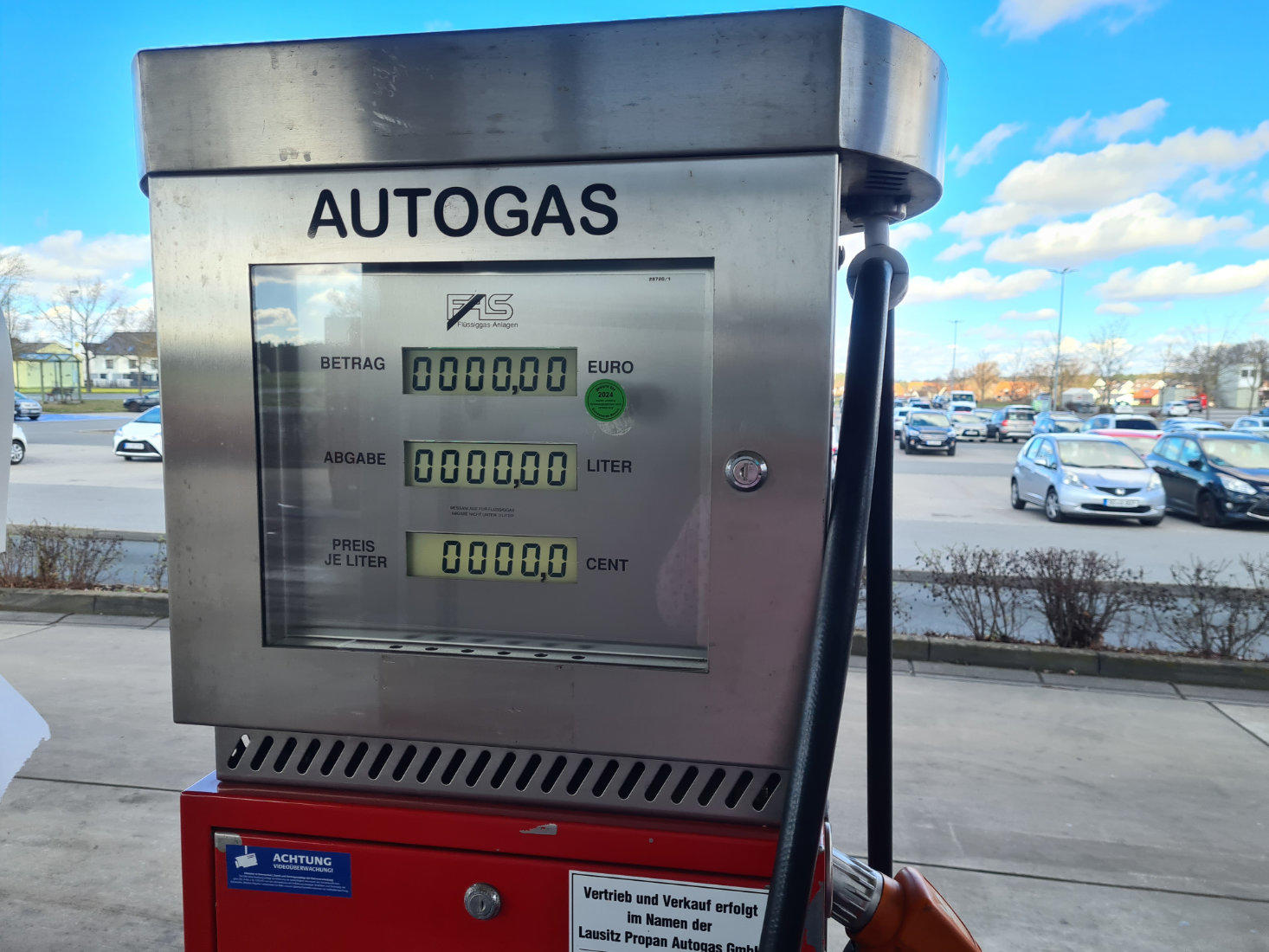 Autogas-Zapfsäule aus Edelstahl mit Aufschrift "AUTOGAS", digitale Anzeige mit Nullen, schwarzer Zapfschlauch rechts, roter Unterbau, Parkplatz mit geparkten Autos und bewölkter Himmel im Hintergrund