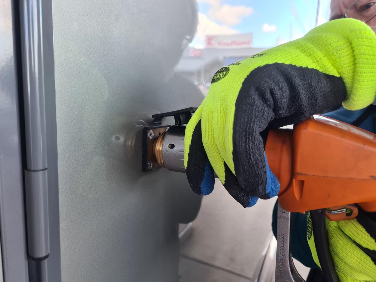 Hand mit neongelbem Arbeitshandschuh hält ein oranges Elektrowerkzeug an einer runden Metalleinheit auf einer grauen Metallfläche