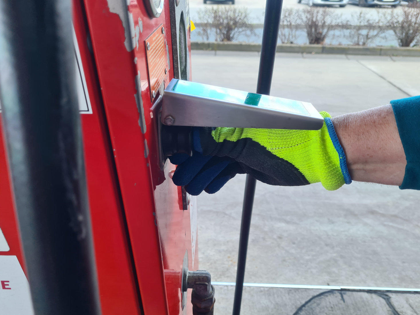 Hand mit gelb-grün-blauem Handschuh hält silbernen Zapfhahn an roter Zapfsäule; schwarzer Schlauch, Asphalt im Hintergrund