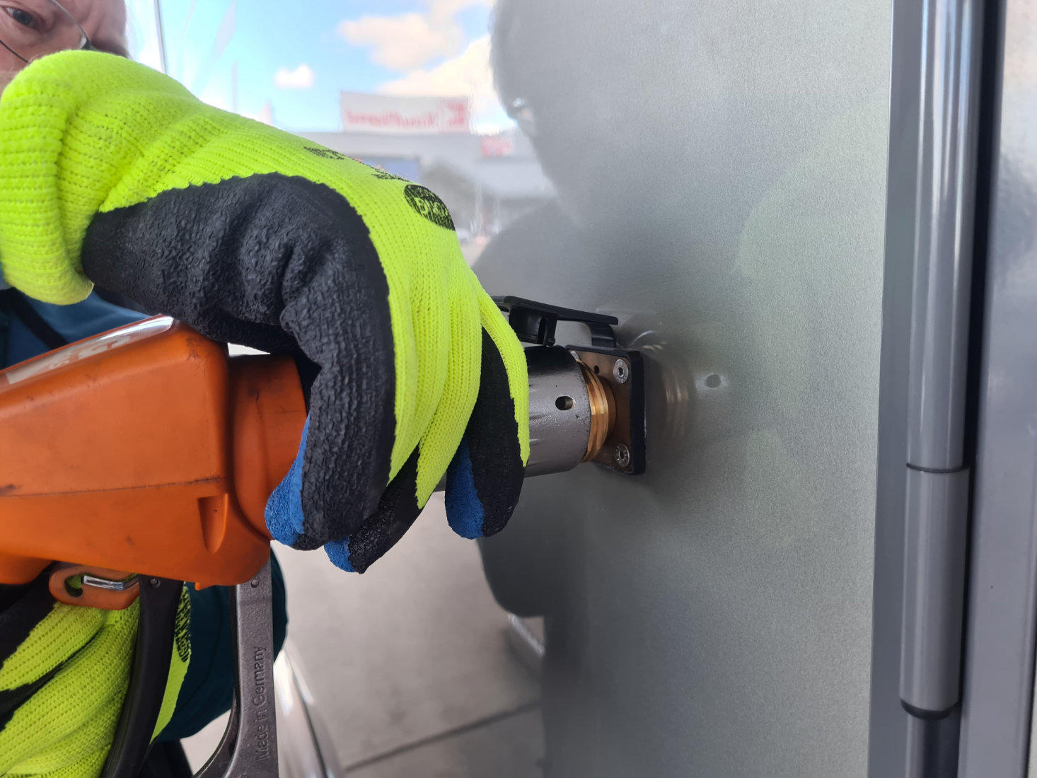 Hand mit neon-gelbem und schwarzem Arbeitshandschuh hält orangefarbene Zapfpistole in runder Öffnung einer grauen Metallfläche