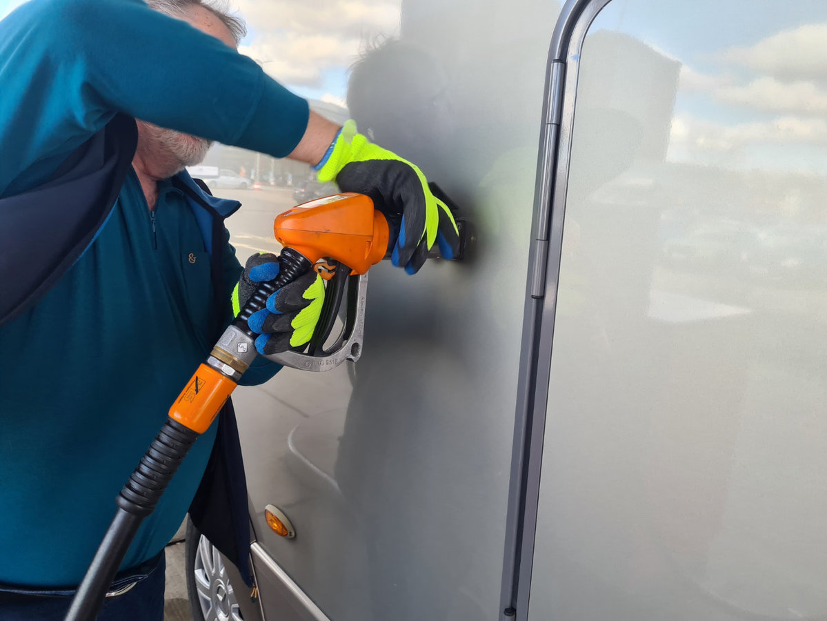 Person mit gelben Handschuhen h&auml;lt eine orangefarbene Zapfpistole mit schwarzem Kabel an die Au&szlig;enseite eines grauen Fahrzeugs