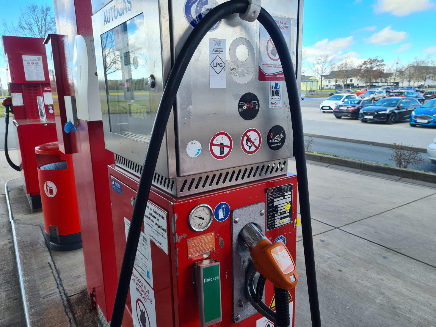 Rote Zapfsäule mit orangefarbener Zapfpistole, Schläuche und Aufkleber; im Hintergrund geparkte Autos und blauer Himmel