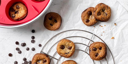 Mehrere kleine runde Kekse mit Schokoladenst&uuml;ckchen auf zerknittertem Backpapier; einige auf einem runden Metallgitter, andere neben einem roten Silikon-Muffinblech; verstreute Schokoladenst&uuml;ckchen und Kr&uuml;mel