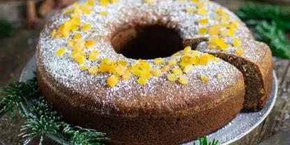 Ringf&ouml;rmiger Kuchen mit einem fehlenden St&uuml;ck, bestreut mit Puderzucker und belegt mit gelben Bl&uuml;tenbl&auml;ttern, auf einem Teller, umgeben von gr&uuml;nen Nadelzweigen