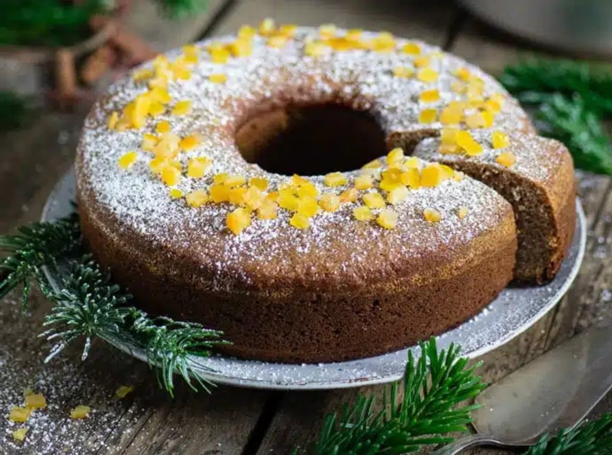 Ringf&ouml;rmiger Kuchen mit einem fehlenden St&uuml;ck, bestreut mit Puderzucker und belegt mit gelben Bl&uuml;tenbl&auml;ttern, auf einem Teller, umgeben von gr&uuml;nen Nadelzweigen