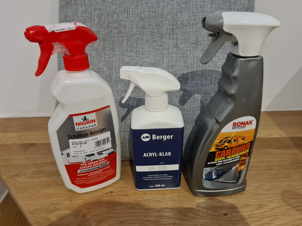 Drei Spr&uuml;hflaschen nebeneinander auf einer Holzplatte vor einem grauen Stoffhintergrund: links wei&szlig;e Flasche mit rotem Spr&uuml;hkopf, Mitte wei&szlig;e Flasche mit blauem Etikett und wei&szlig;em Spr&uuml;hkopf, rechts graue Flasche mit wei&szlig;em Spr&uuml;hkopf und gelbem Etiket