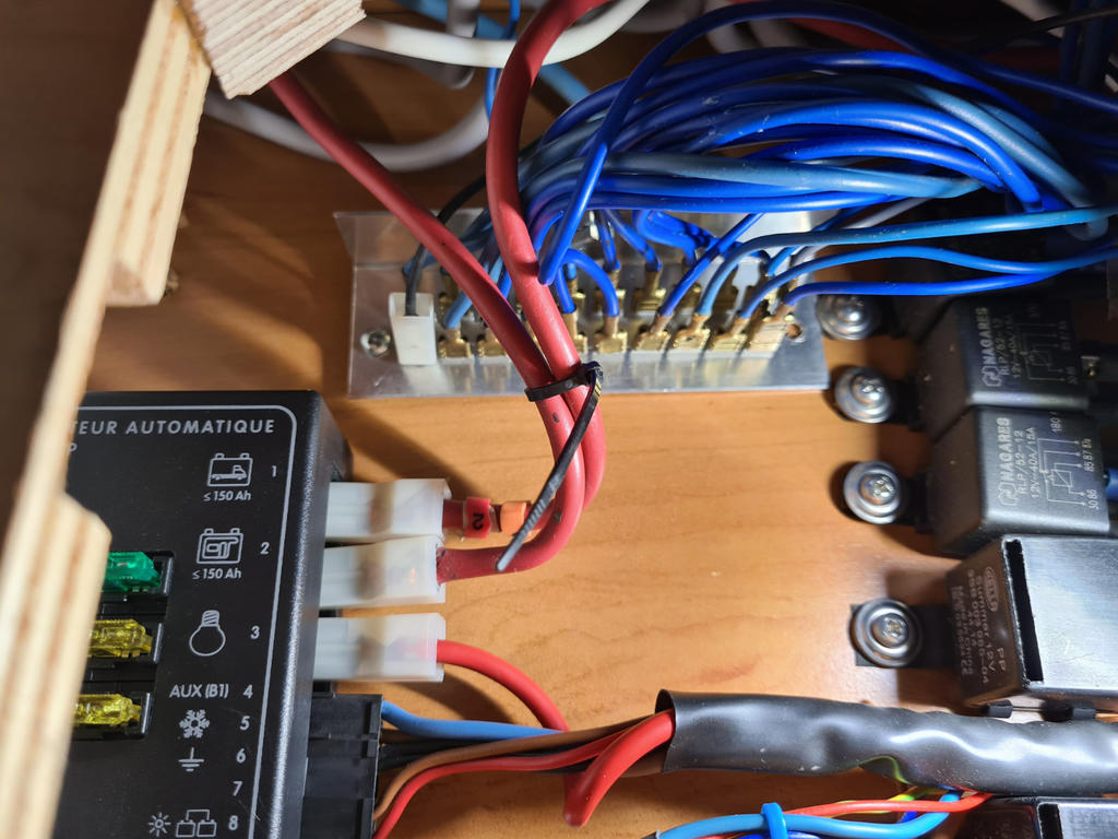 Innenraum eines Holzgehäuses mit blauen, roten und schwarzen Kabeln, weißer Steckverbindung, Kontaktleiste mit goldfarbenen Stiften, drei schwarzen rechteckigen Bauteilen, Schrauben und Kabelbinder