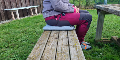 Eine sitzende Person in violett-grauer Jacke und dunkelroten Hosen auf einem grauen Sitzkissen auf einer h&ouml;lzernen Picknickbank; weitere Holztische und -b&auml;nke, Gras, Zaun und Vegetation im Hintergrund