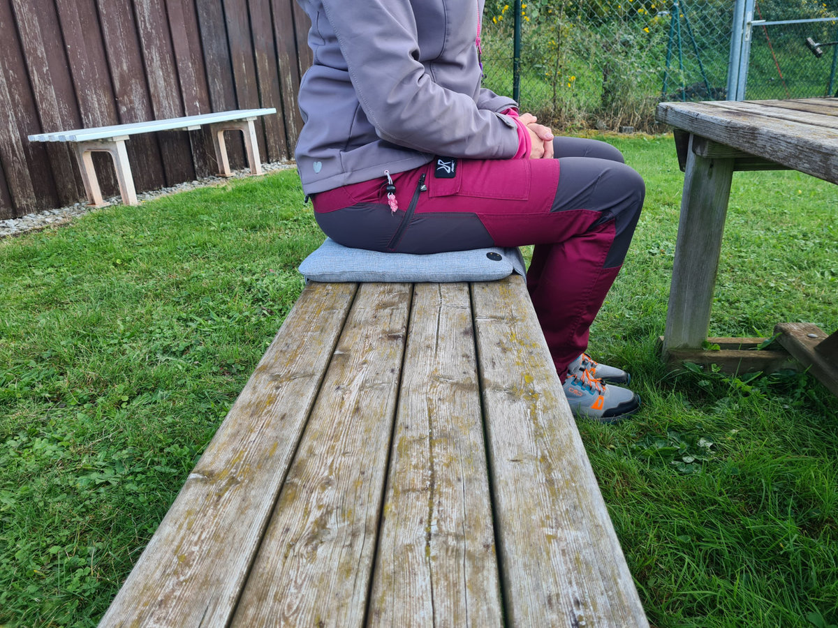 Eine sitzende Person in violett-grauer Jacke und dunkelroten Hosen auf einem grauen Sitzkissen auf einer h&ouml;lzernen Picknickbank; weitere Holztische und -b&auml;nke, Gras, Zaun und Vegetation im Hintergrund