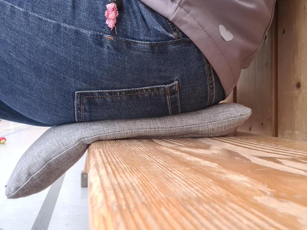 Unterer R&uuml;cken und Ges&auml;&szlig; einer sitzenden Person auf einer Holzbank mit grauem Sitzkissen; blaue Jeans mit rosa Blumenanh&auml;nger, graue Jacke