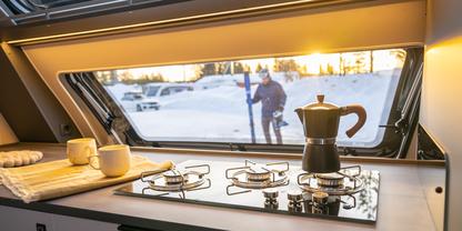 Innenraum eines Wohnmobils mit Gasherd, Espressokanne, Tassen und Handtuch; durch das Fenster: verschneite Landschaft und eine Person mit Skiern