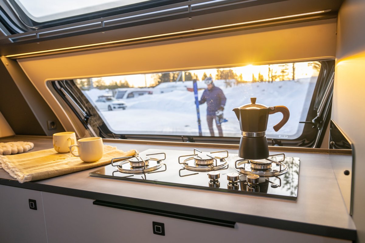 Innenraum eines Wohnmobils mit Gasherd, Espressokanne, Tassen und Handtuch; durch das Fenster: verschneite Landschaft und eine Person mit Skiern
