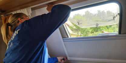 Person mit blondem Zopf und blauem Oberteil h&auml;lt den Rahmen eines ge&ouml;ffneten Dachfensters &uuml;ber einem gepolsterten Sitz in einem Innenraum