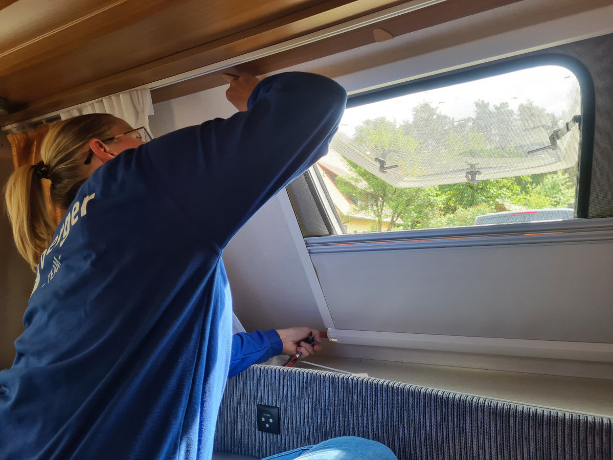 Person mit blondem Zopf und blauem Oberteil h&auml;lt den Rahmen eines ge&ouml;ffneten Dachfensters &uuml;ber einem gepolsterten Sitz in einem Innenraum